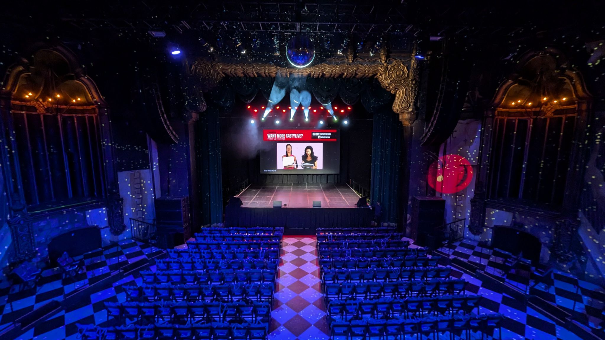An ornate theater venue with stage lighting, a large tastylive screen, and rows of seats on a checkered floor